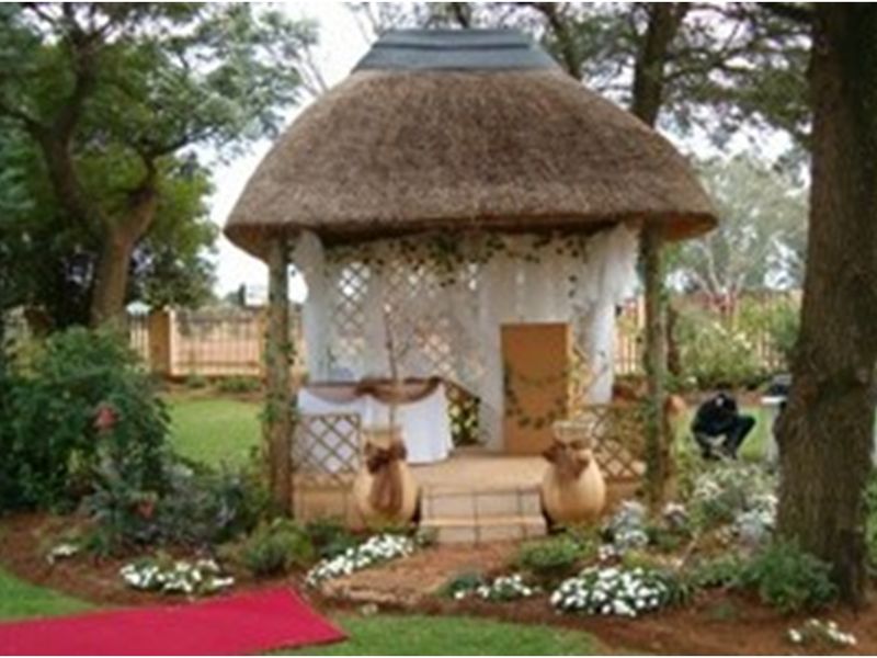 A charming thatched roof gazebo set within a landscaped garden, featuring timber decking and lattice screening, ideal for outdoor events or ceremonies.