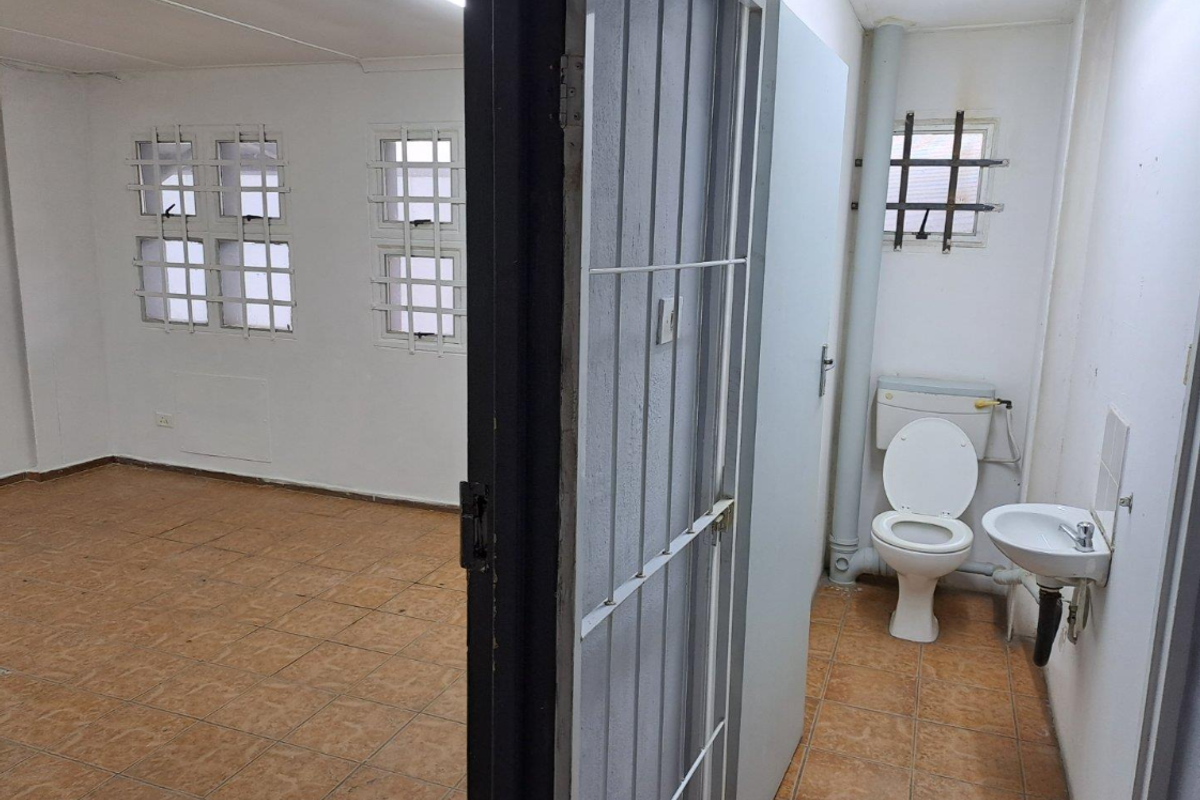 An industrial-style interior space featuring a tiled room adjacent to a small, functional bathroom with basic sanitary fixtures and security bars on windows.