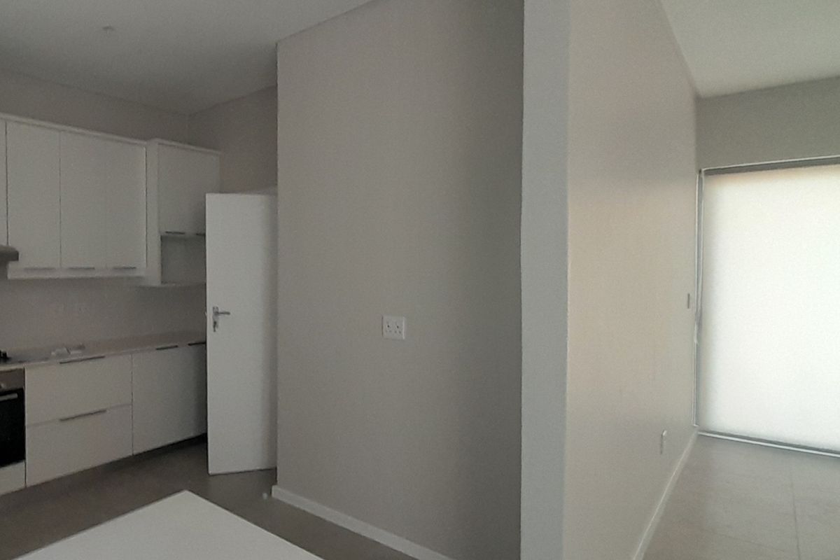 A modern open-plan kitchen area featuring white cabinetry, integrated oven, and tiled flooring, leading to a bright living space with sliding glass doors.