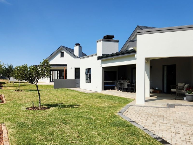 A well-maintained rear exterior view featuring a covered patio with a built-in braai, paved walkways, and a neatly landscaped garden.