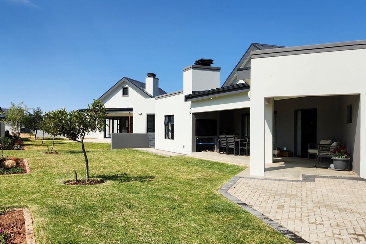 A well-maintained rear exterior view featuring a covered patio with a built-in braai, paved walkways, and a neatly landscaped garden.
