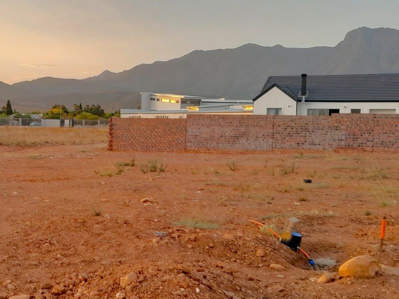 A vacant residential plot featuring a brick boundary wall and a water connection point, set against a scenic mountain backdrop.