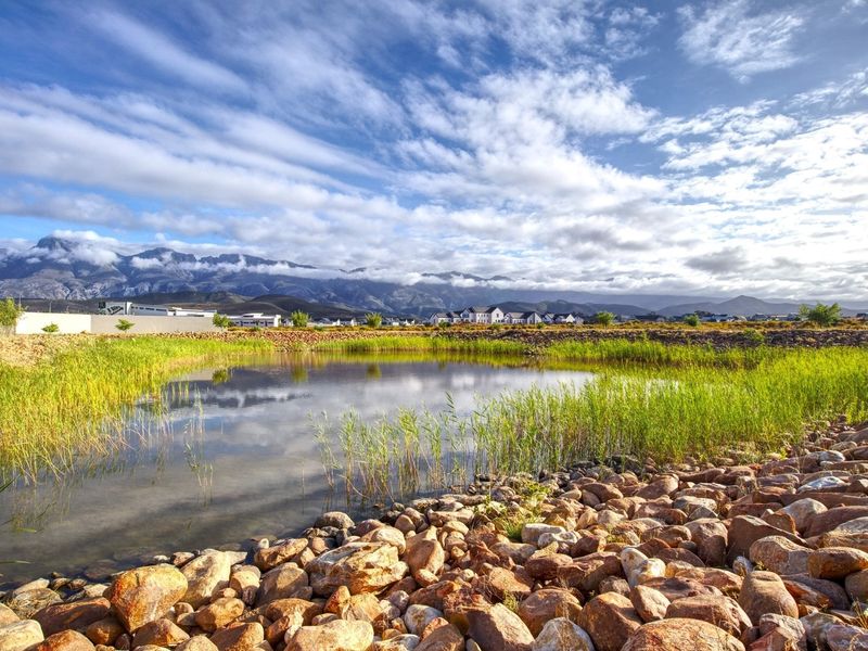 A scenic view of a natural pond with rock landscaping and reeds, set against a backdrop of mountains and a clear sky.