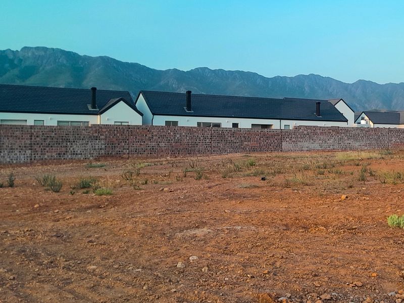 A view of a vacant residential plot featuring a brick boundary wall with neighboring houses and mountain views in the background.