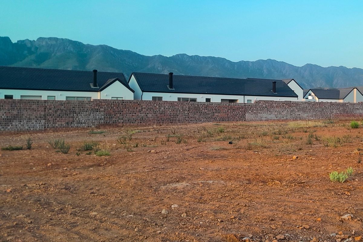A view of a vacant residential plot featuring a brick boundary wall with neighboring houses and mountain views in the background.