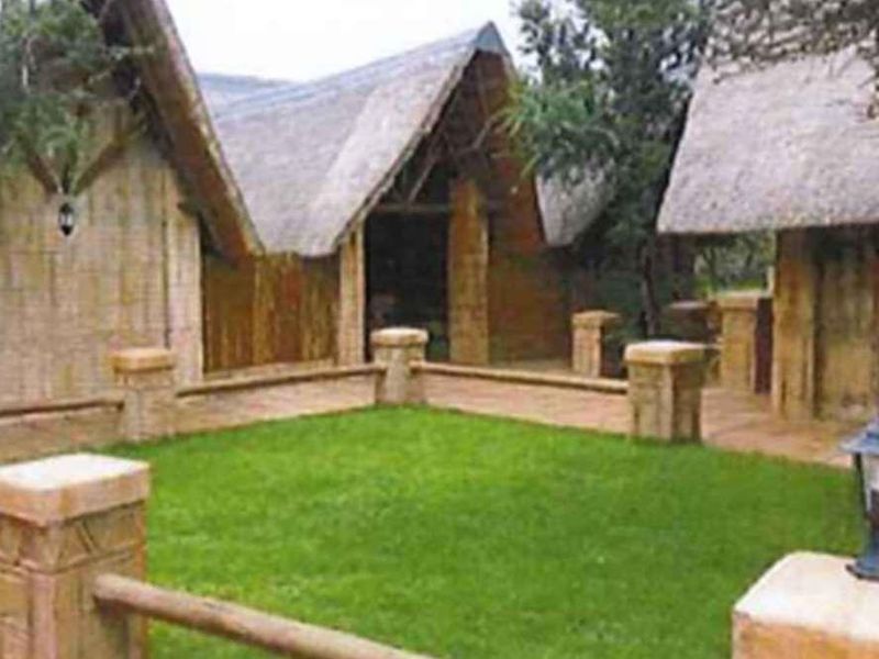 A rustic exterior view of a small holding featuring traditional thatched roof structures, a central lawn, and stone-pillar fencing.