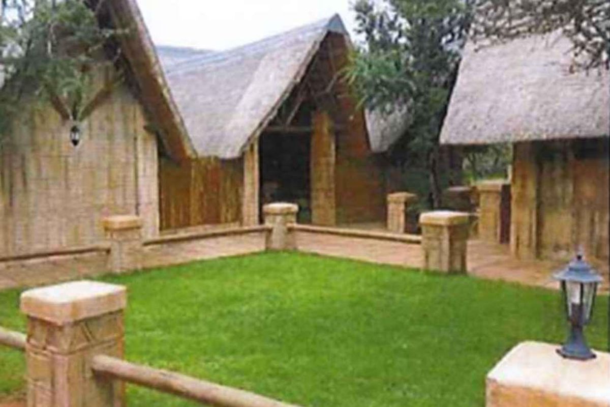 A rustic exterior view of a small holding featuring traditional thatched roof structures, a central lawn, and stone-pillar fencing.
