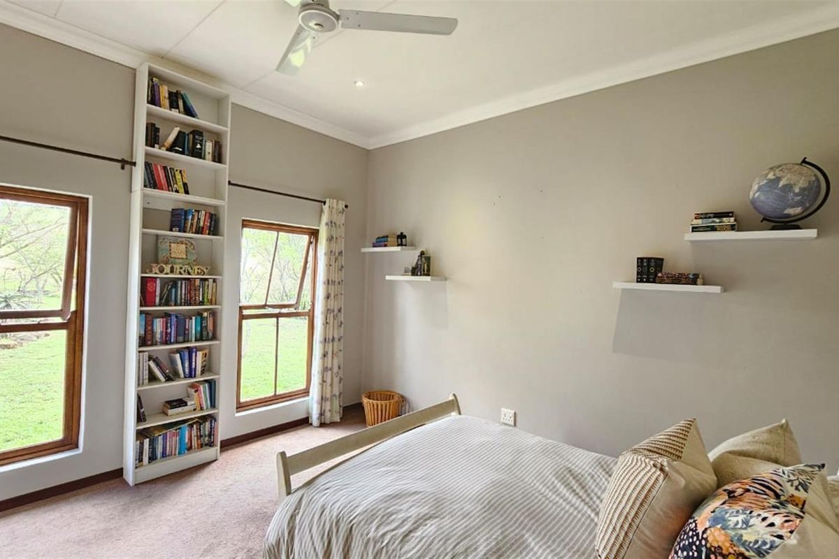 A well-lit bedroom featuring a built-in floor-to-ceiling bookshelf, ceiling fan, and large casement windows overlooking the garden.