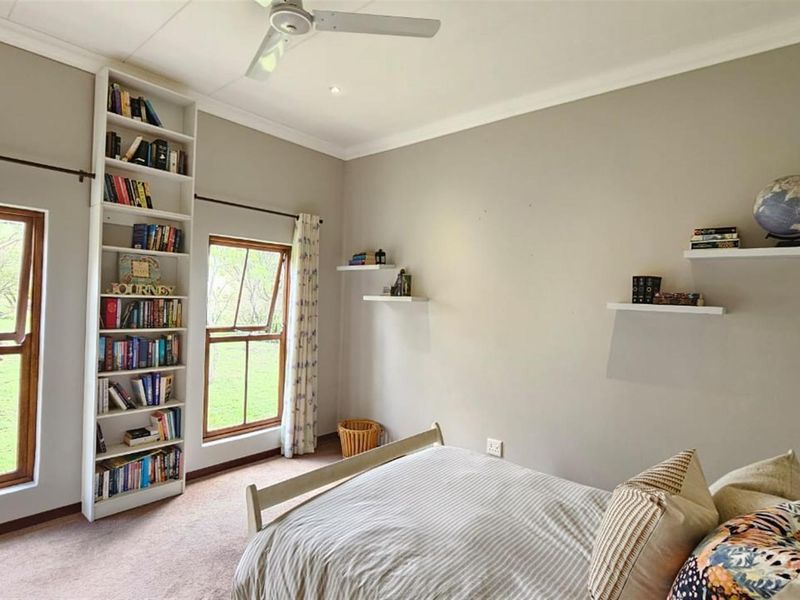 A well-lit bedroom featuring a built-in floor-to-ceiling bookshelf, ceiling fan, and large casement windows overlooking the garden.