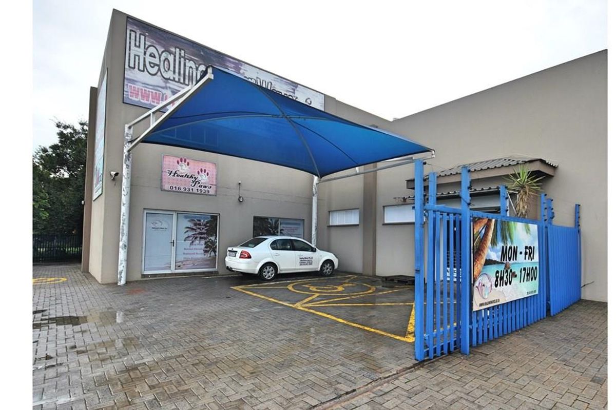 Commercial property exterior featuring a paved parking area, blue shade sail structure, and secure gated entrance with business signage.