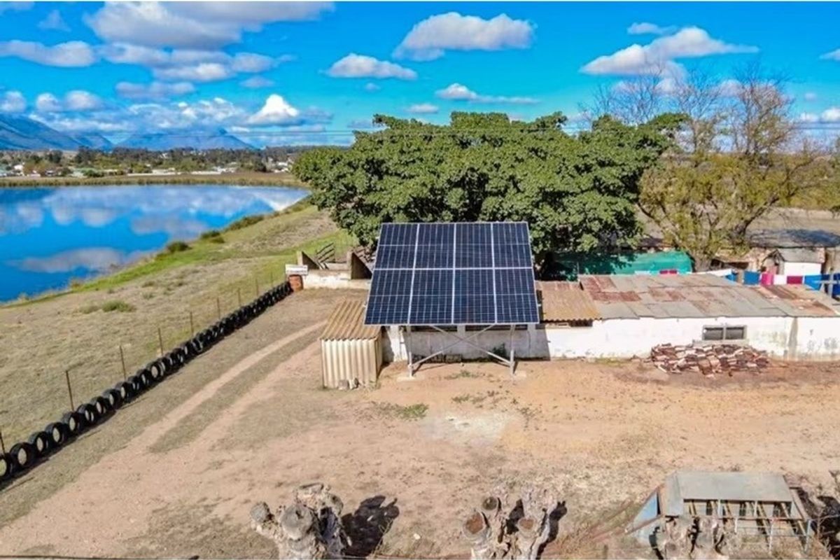 Aerial view of a small holding featuring a ground-mounted solar panel array, adjacent outbuilding, and scenic dam frontage.