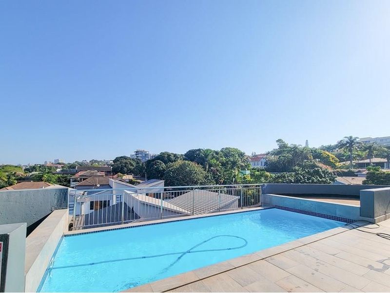 A rooftop swimming pool area featuring a tiled deck, metal safety balustrades, and panoramic views of the surrounding neighborhood.