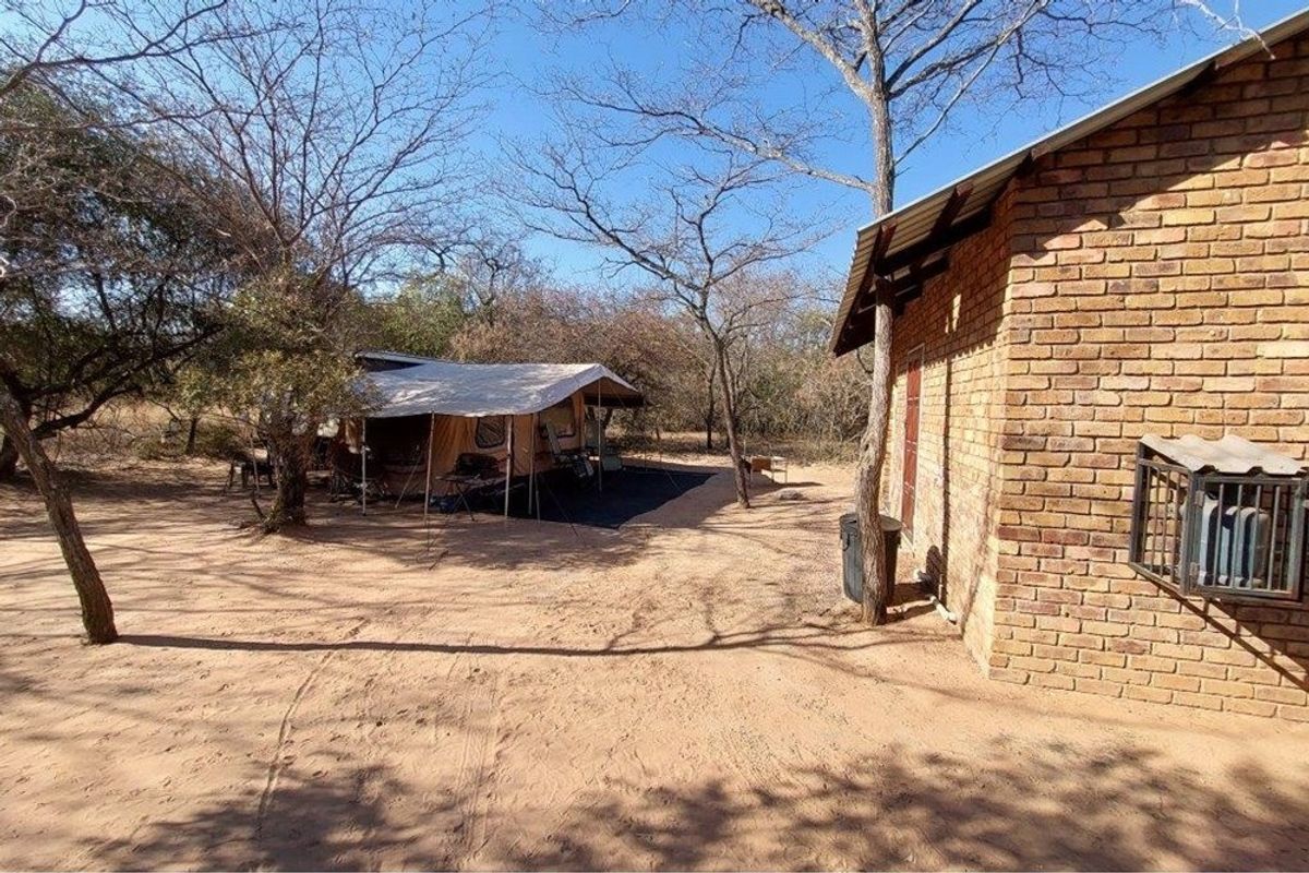 A rustic farm setting featuring a brick structure and a canvas tent setup nestled among natural trees and sandy terrain.