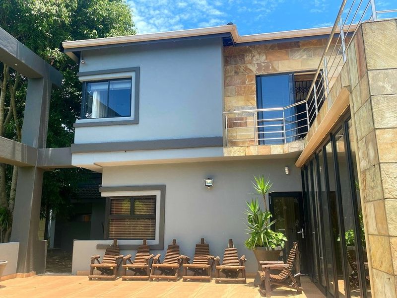 Rear elevation of a modern two-story house featuring stone cladding, a spacious tiled patio, and a balcony with stainless steel railings.