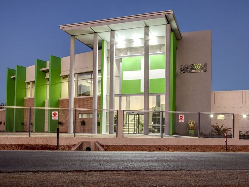 A modern, well-maintained private hospital building facade featuring contemporary architectural design, prominent green accents, and secure perimeter fencing at dusk.