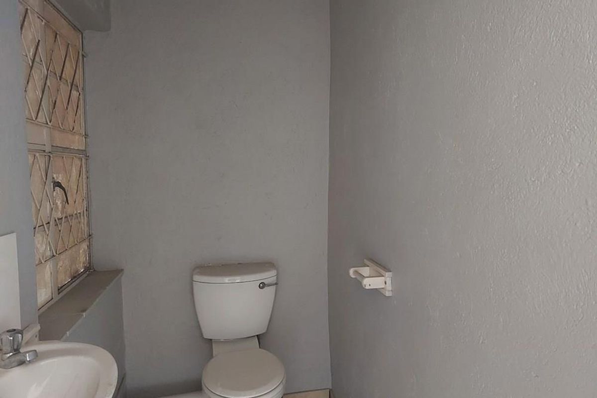 A compact commercial restroom featuring a standard toilet, wall-mounted basin, and a security-style window with metal burglar bars.