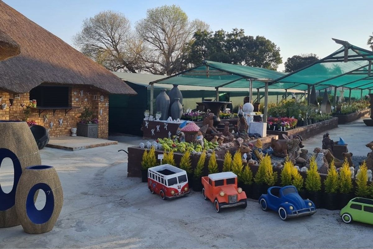 Outdoor retail area featuring a thatched roof structure, paved ground, and shaded display sections for garden ornaments and plants.