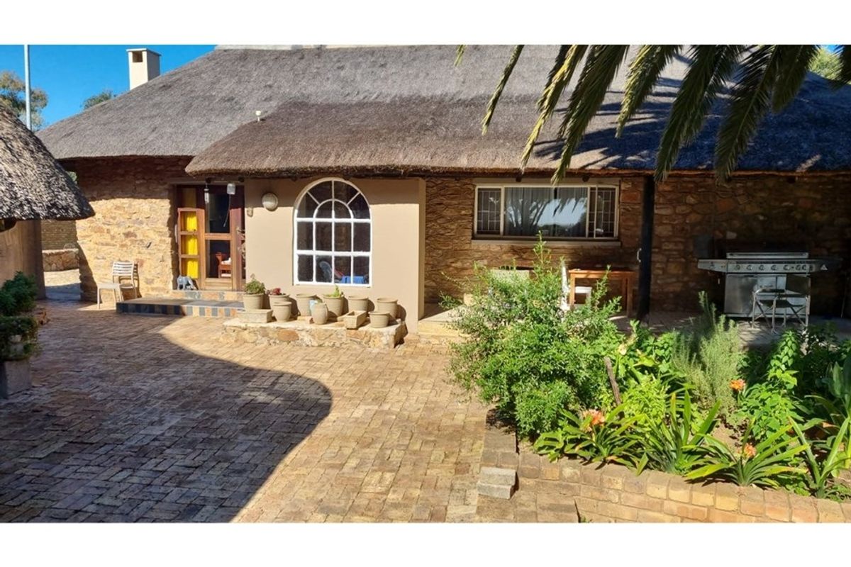 A charming rear exterior view featuring a traditional thatched roof, stone-clad walls, arched window, and a paved patio area with established garden beds.