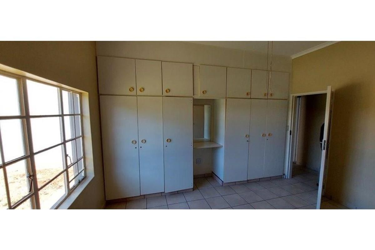 A spacious bedroom featuring extensive built-in cupboards, tiled flooring, and natural light through a large window with security bars.