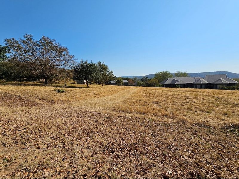 A wide-angle view of a vacant residential plot featuring natural grass, mature trees, and distant views of neighboring properties and hills.