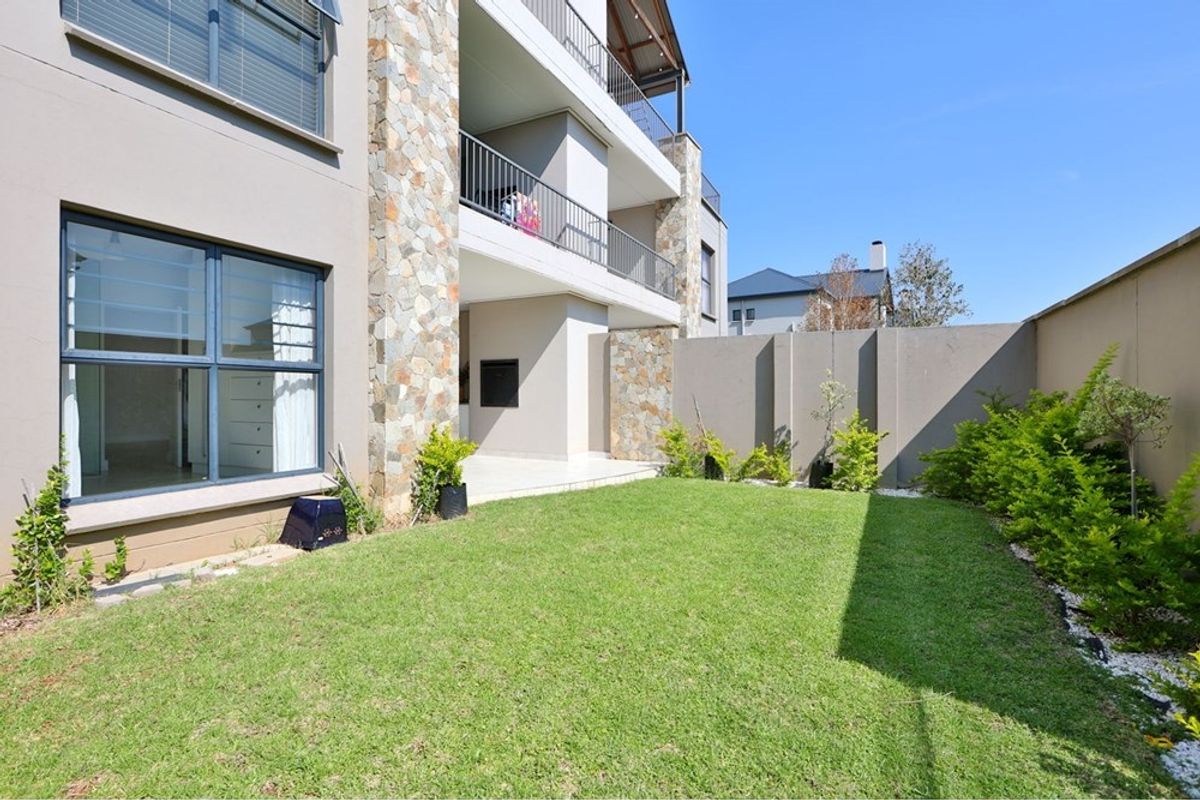 A private rear garden featuring a manicured lawn, a covered patio with a built-in braai, and stone-clad architectural detailing on the apartment building facade.