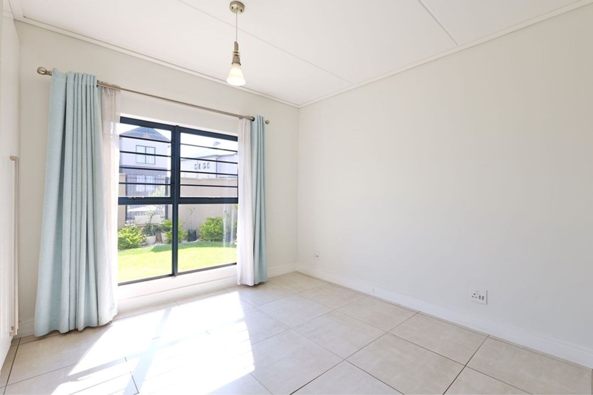 A bright, empty bedroom featuring neutral tiled flooring, a built-in cupboard, and a large window with security bars overlooking the garden.