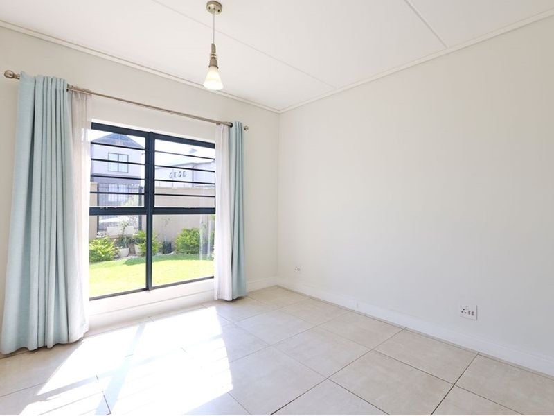 A bright, empty bedroom featuring neutral tiled flooring, a built-in cupboard, and a large window with security bars overlooking the garden.