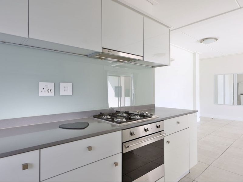 Modern kitchen featuring sleek white cabinetry, stone countertops, a gas hob, built-in oven, and a glass splashback, set within an open-plan living space.