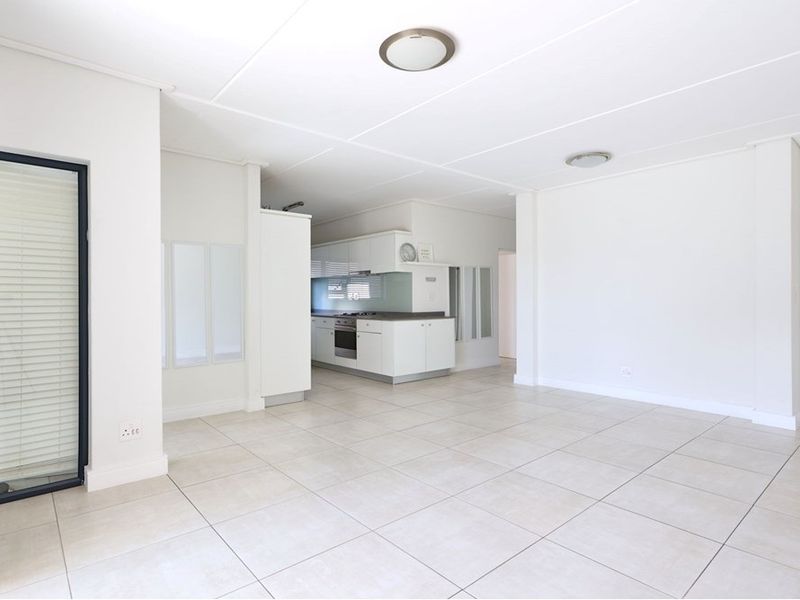 A bright, open-plan living and kitchen area featuring neutral tiled flooring, modern white cabinetry, and integrated kitchen appliances in a clean, vacant space.