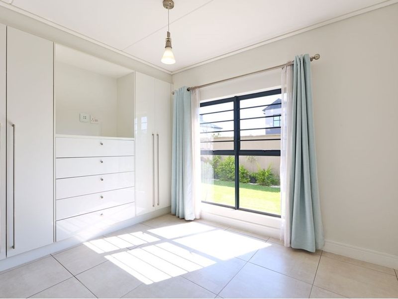 A bright, empty bedroom featuring modern white built-in cupboards, tiled flooring, and a window with security bars overlooking the garden.
