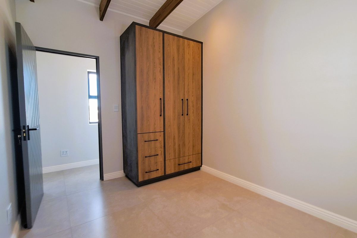 A clean, modern bedroom featuring built-in wooden wardrobes, exposed timber ceiling beams, and neutral tiled flooring, offering a bright and well-maintained living space.