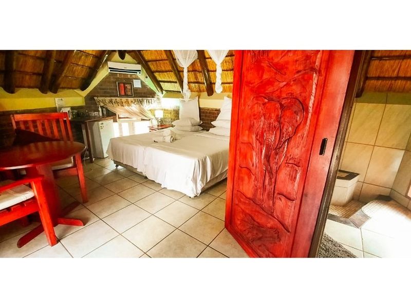 A bedroom in a cottage featuring a traditional thatched roof, exposed brick walls, tiled flooring, and a distinctive carved wooden door.