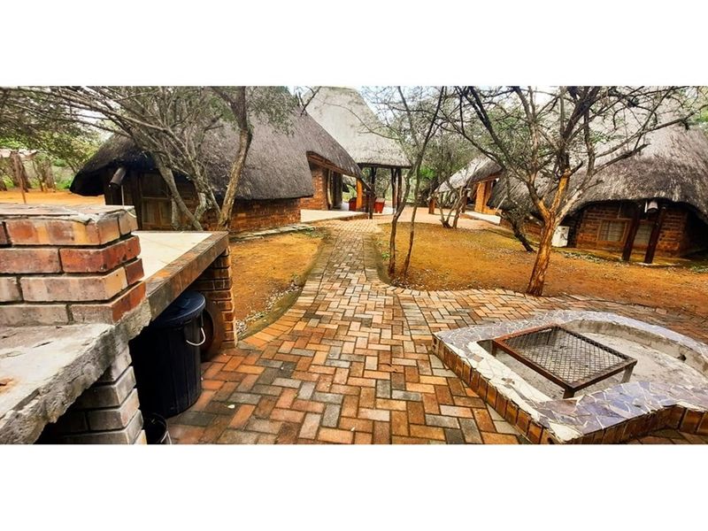 Outdoor entertainment area featuring a brick braai and built-in fire pit, set within a garden space with traditional thatched cottage structures.