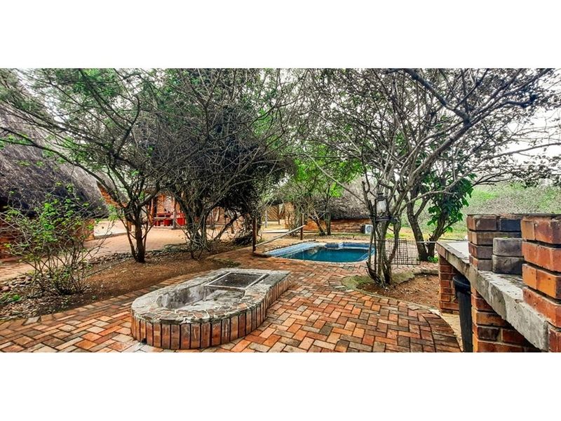 Outdoor entertainment area featuring a swimming pool, brick paved patio, built-in brick braai, and a fire pit surrounded by mature trees and thatched cottages.