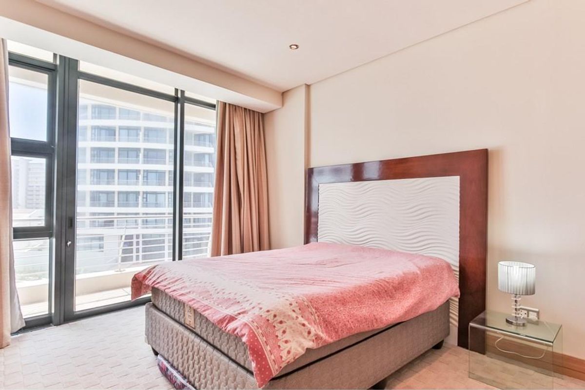 A bright bedroom featuring floor-to-ceiling windows with balcony access, neutral carpeting, and a built-in headboard design.