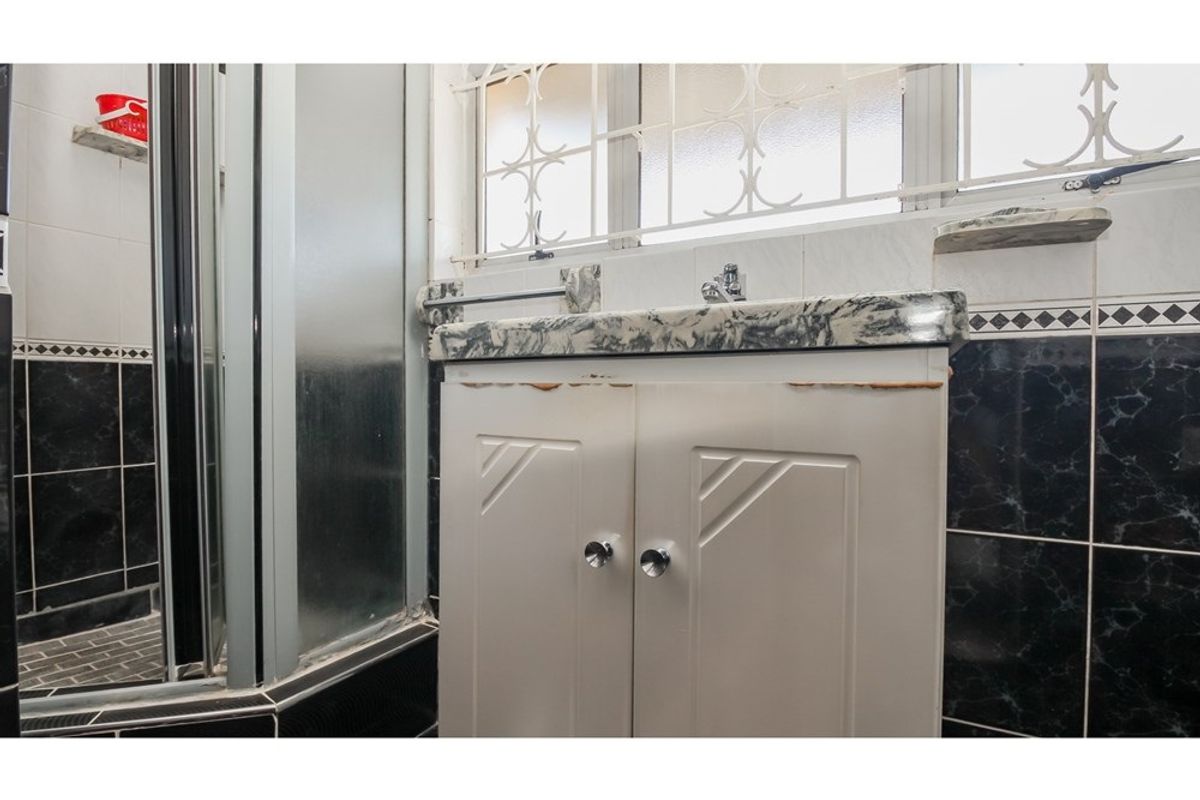 A compact bathroom featuring a vanity unit with a marble-effect countertop, a shower enclosure, and decorative wall tiling with security bars on the window.