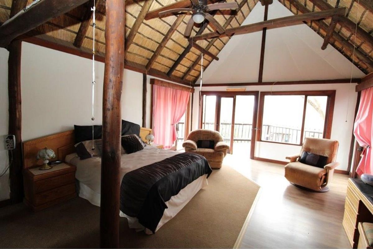 Spacious bedroom featuring a high vaulted thatch ceiling with exposed timber beams, wooden support pillars, and large glass sliding doors leading to a balcony.