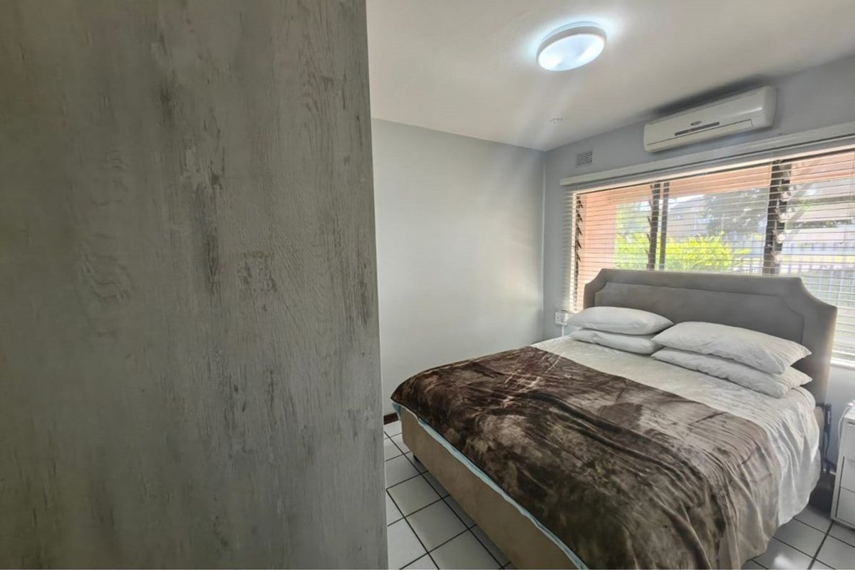 A bedroom featuring tiled flooring, a wall-mounted split-system air conditioner, and louvre windows providing natural light.
