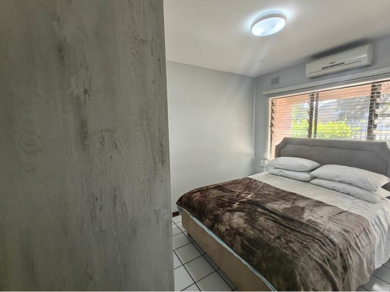A bedroom featuring tiled flooring, a wall-mounted split-system air conditioner, and louvre windows providing natural light.