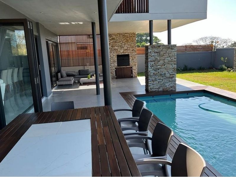 Spacious covered patio featuring a built-in braai, stone cladding accents, and seamless flow to a timber-decked swimming pool and garden area.