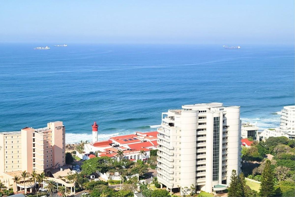An elevated aerial view showcasing the coastal apartment complex, surrounding buildings, and the expansive ocean horizon.