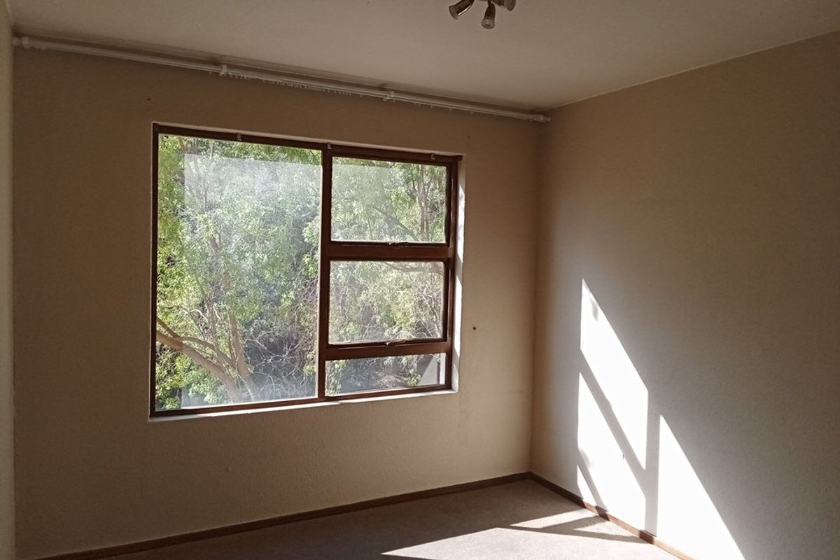An empty bedroom featuring a large window with a leafy outlook, carpeted flooring, and surface-mounted electrical conduit along the ceiling line.