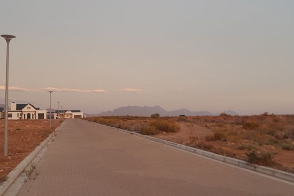 A view of a paved residential street with concrete curbing and street lighting, set against a backdrop of open land and distant mountains.