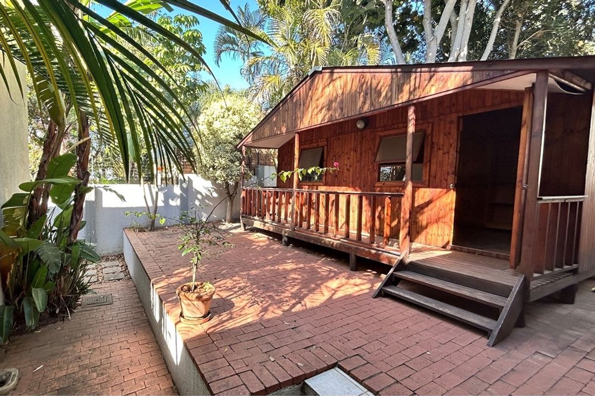 A detached wooden cabin featuring a covered front veranda and brick-paved patio area, set within a landscaped garden environment.