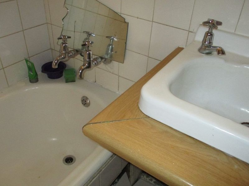 A dated bathroom featuring a bathtub with wall-mounted taps and a vanity basin with a damaged countertop and cracked mirror.