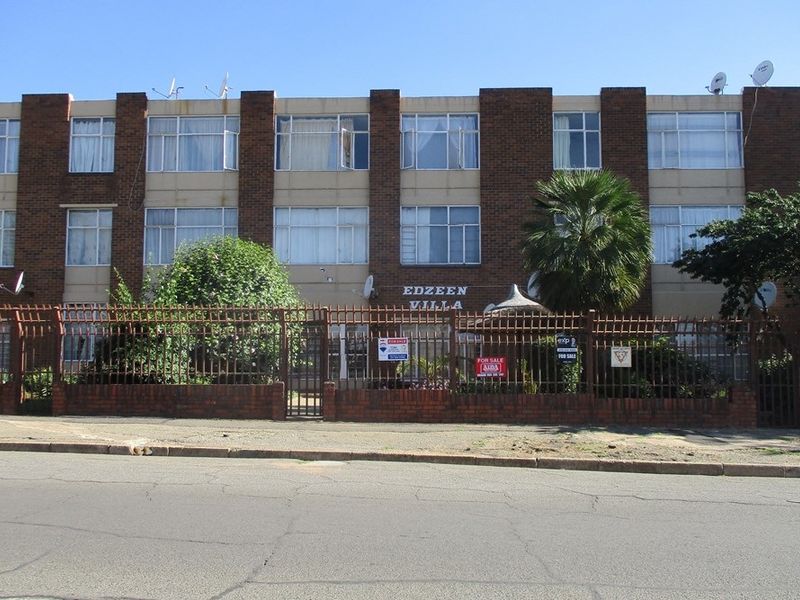 Front elevation of Edzeen Villa apartment complex featuring a brick facade, secure perimeter fencing, and established landscaping.