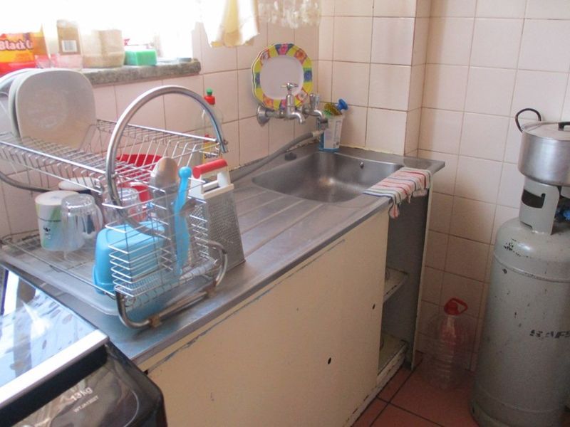 A compact kitchen area featuring a stainless steel sink, wall-mounted taps, and tiled splashbacks, requiring modernization of finishes and cabinetry.