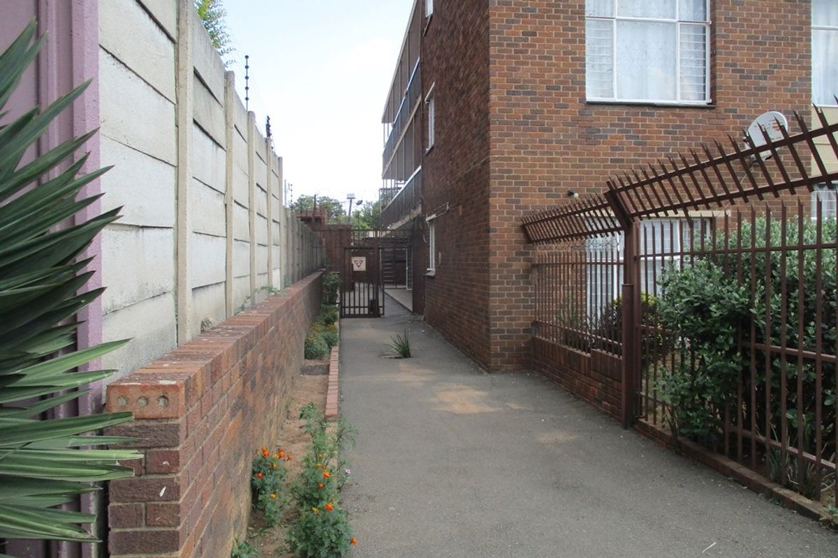 A narrow paved walkway between a brick apartment building and a boundary wall, featuring security fencing and a metal access gate.