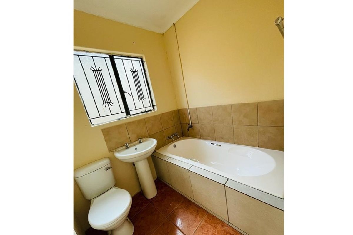 A functional bathroom featuring a built-in bathtub, pedestal basin, and toilet with tiled splashbacks and security bars on the window.