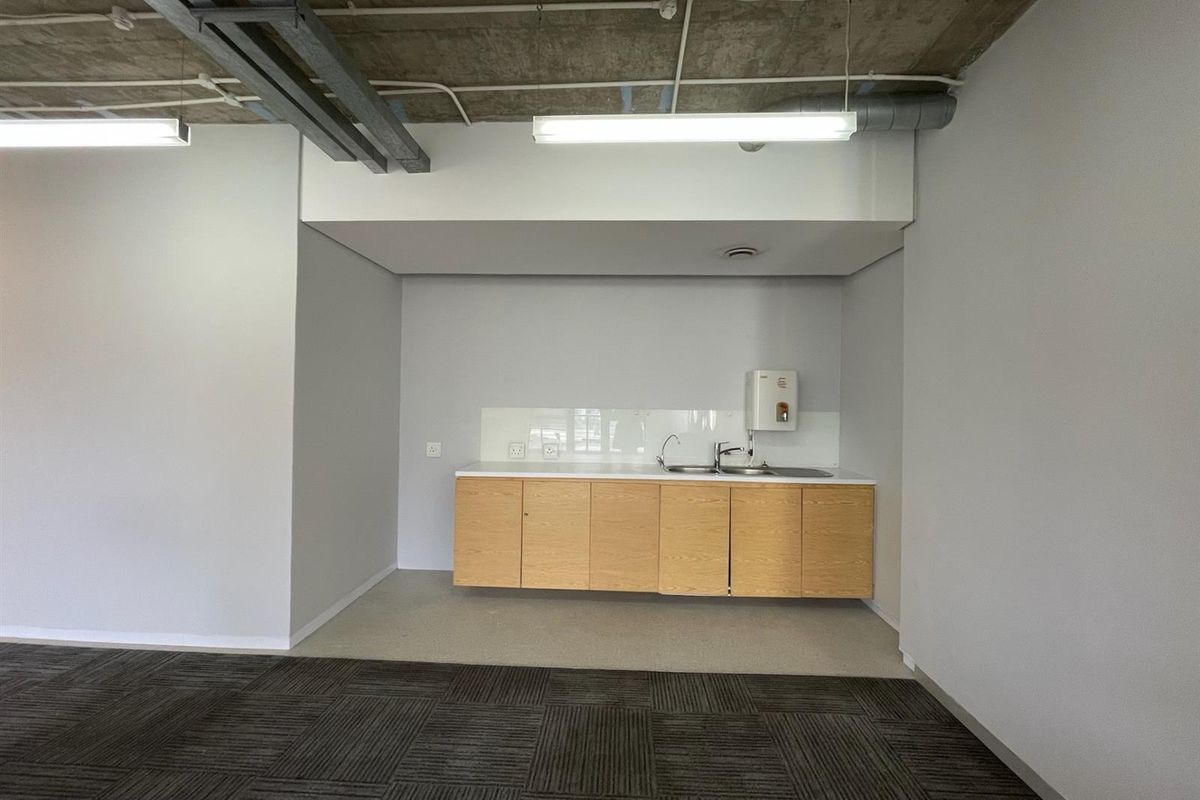 A clean, modern office kitchenette featuring light wood cabinetry, a stainless steel sink, and an exposed industrial-style concrete ceiling.